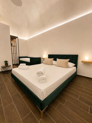 Una cama bien hecha con cabecera verde y ropa de cama blanca, una mesita de noche con una lámpara y una planta en maceta sobre una mesa redonda.