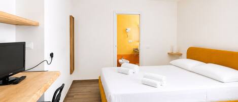 A hotel room with a bed, a desk with a computer, a TV, and a bathroom visible through an open door.