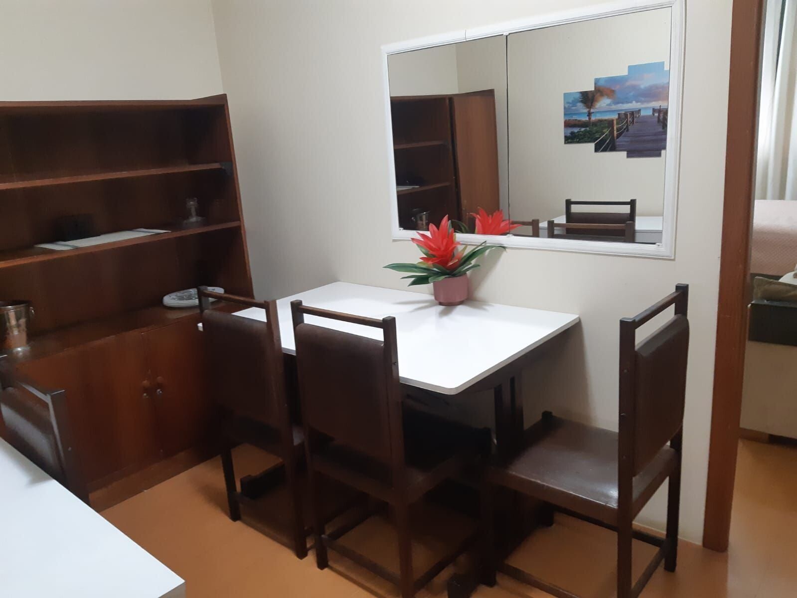 Una habitación pequeña con un escritorio blanco, dos sillas, una estantería y un espejo en la pared.