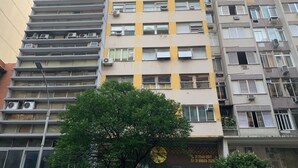 A multi-story residential building with air conditioning units on each floor, a visible door with contact numbers, and a tree in the foreground. - Ap Copacabana 200m da praia - CP5 (Rio de Janeiro)