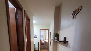 A narrow hallway with wooden flooring and white walls. There is a door on the left, a shelf with a potted plant on the right, and a glimpse of a room with a bed and a window in the distance. - Ap Copacabana 200m da praia - CP5 (Rio de Janeiro)