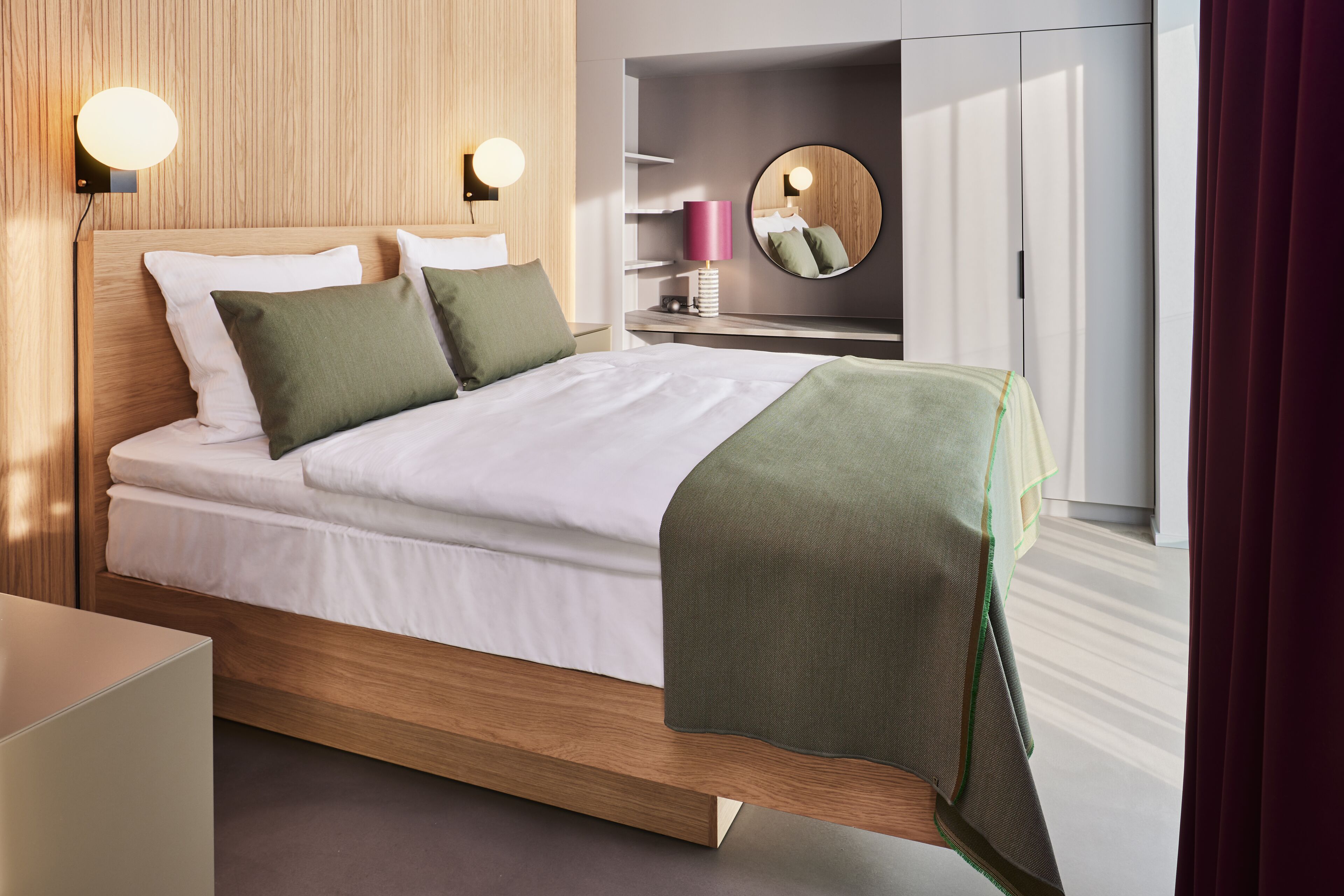 Una habitación de hotel moderna con cabecera de cama de madera, ropa de cama blanca y un plaid verde. Se observa una mesita de noche con una lámpara y un espejo encima.