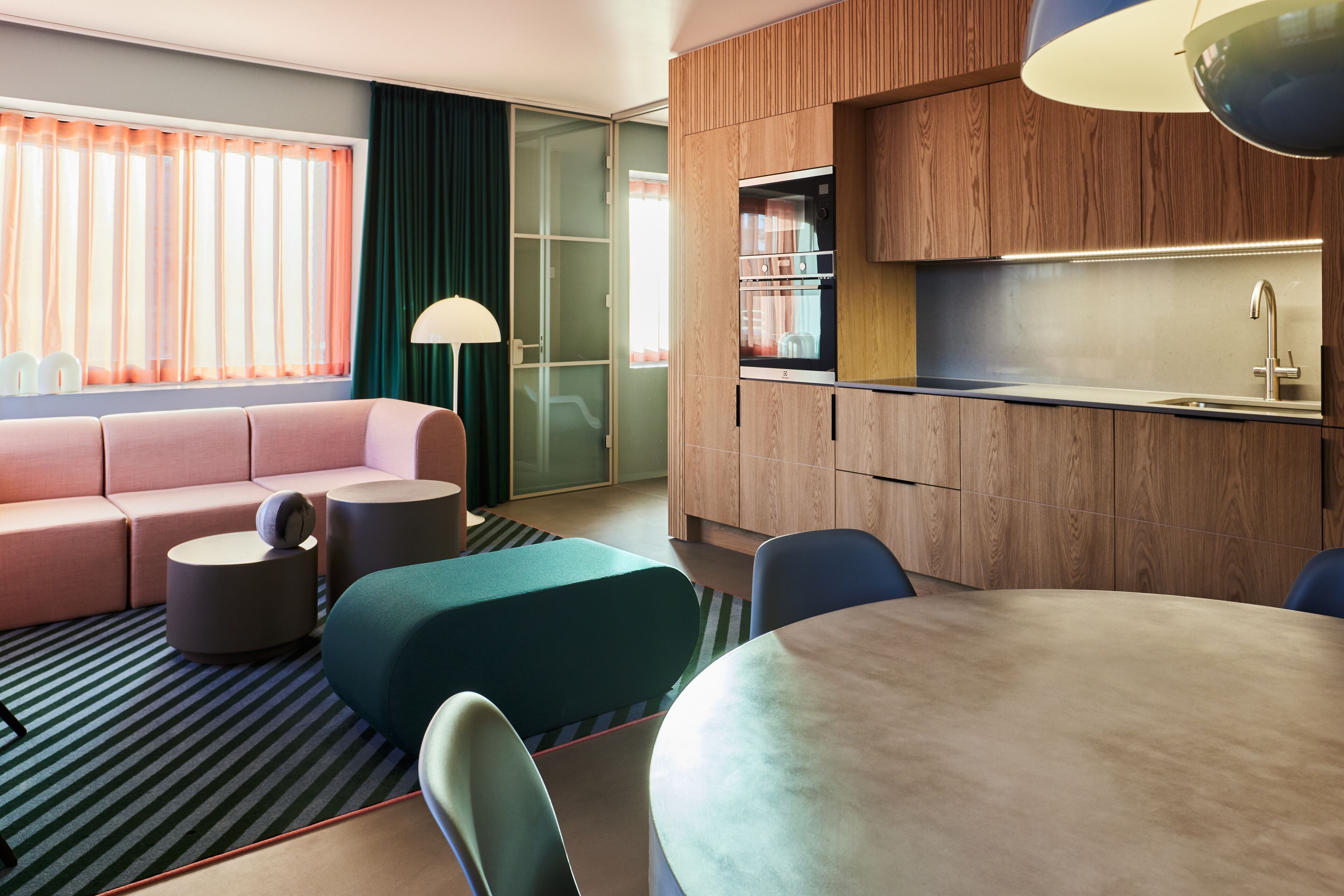 A modern hotel room with a sofa, ottoman, round table, chairs, a kitchen area, and a large window with curtains.