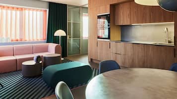 A modern hotel room with a sofa, ottoman, round table, chairs, a kitchen area, and a large window with curtains.