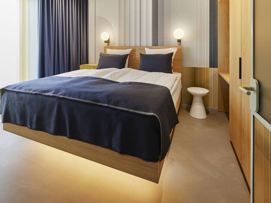 Quarto de hotel moderno com uma cama grande, mesas de cabeceira, uma mesinha de cabeceira menor, um guarda-roupa de madeira e um toalete branco.