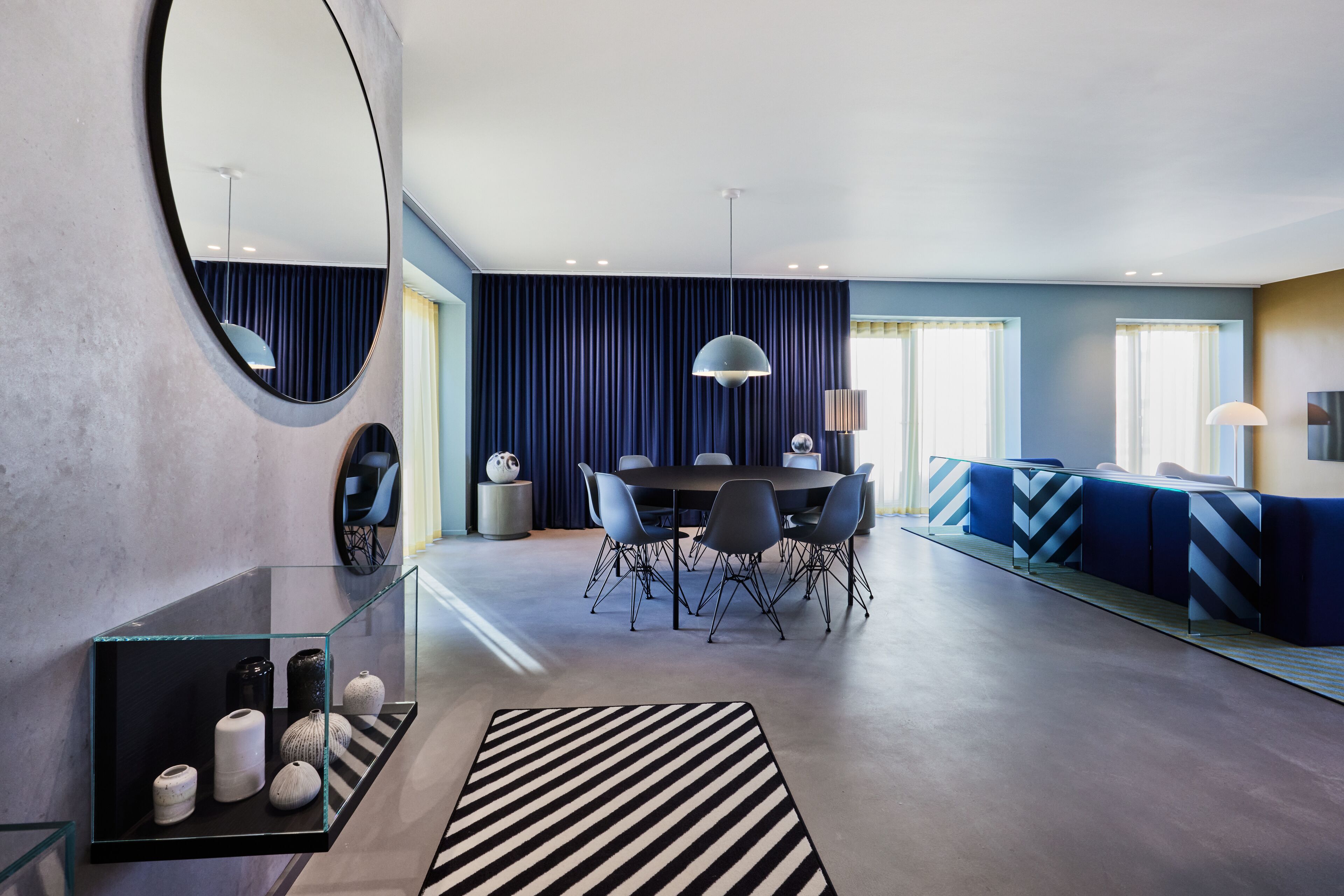 A modern living room with a dining area, featuring a round mirror, a glass coffee table, and a striped rug.