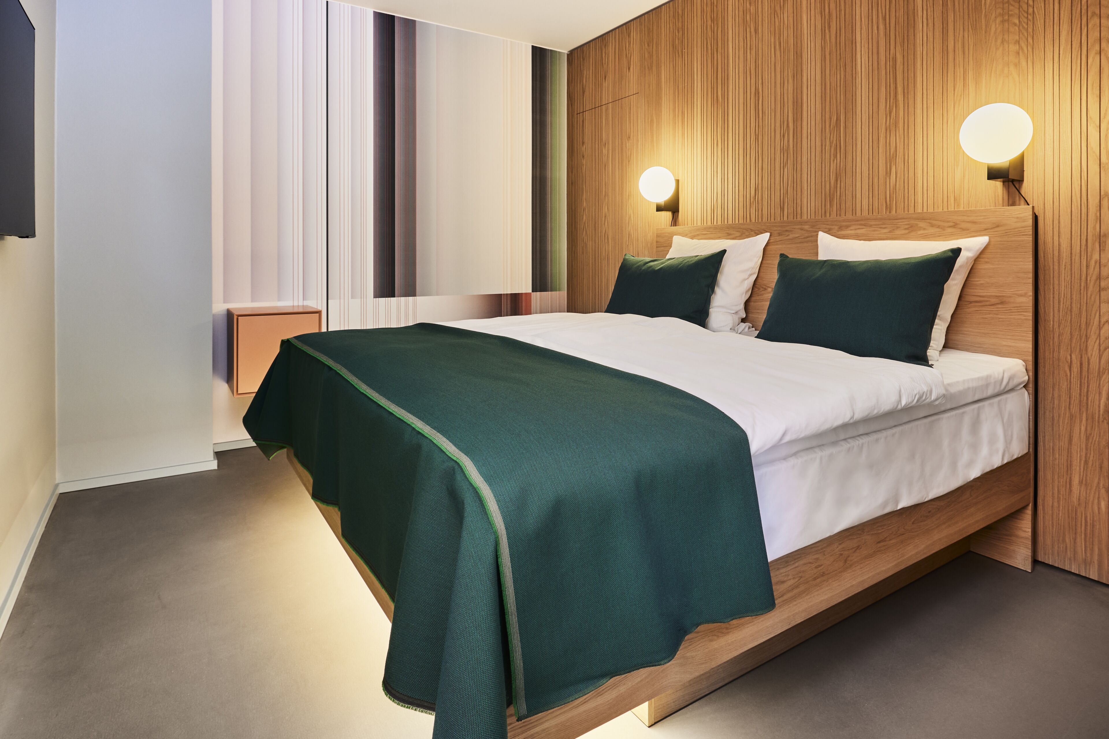A modern hotel room with a wooden headboard, a green bedspread, and two wall-mounted lights.