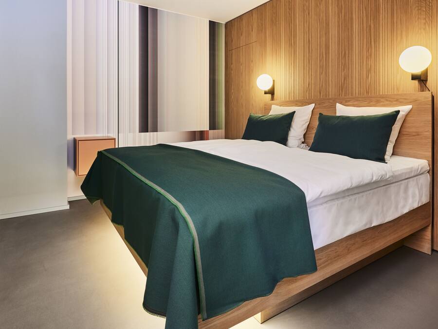 A modern hotel room with a wooden headboard, a green bedspread, and two wall-mounted lights.