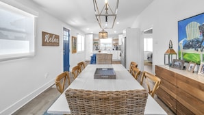 Une cuisine moderne avec des meubles blancs, un coin repas avec une table blanche et des chaises en osier, et un luminaire suspendu.