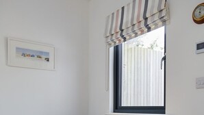 Une pièce avec une table en bois, deux chaises blanches, une fenêtre avec des rideaux à rayures, un tableau encadré au mur et une horloge.