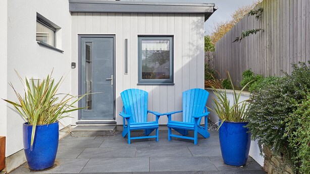 Une maison moderne avec une façade grise, une porte d’entrée et deux fenêtres. On y trouve deux chaises bleues et deux grands pots de plantes.