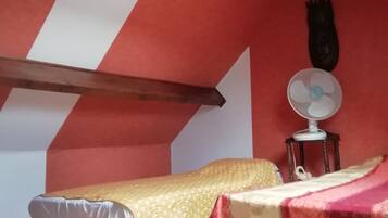 A small, cozy room with a sloped ceiling, a bed with a yellow bedspread, a bedside table with a fan, and a red and white striped wall.