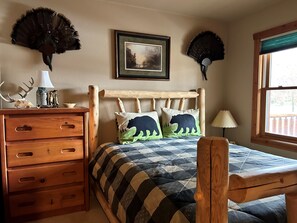 Un dormitorio con estructura de cama de madera, un aparador con cajones, un cuadro de paisaje enmarcado y dos ventiladores de pared.