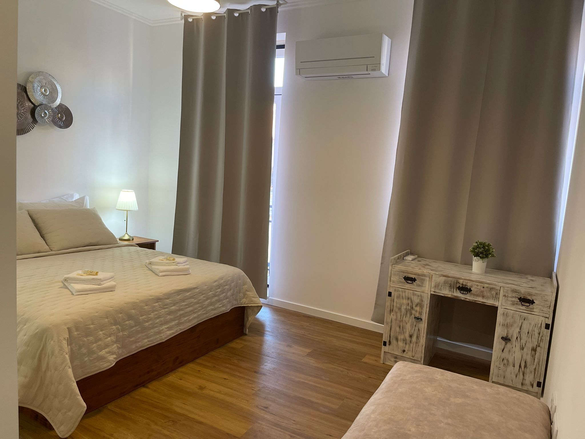 A hotel room with a bed, bedside lamp, air conditioning unit, wooden nightstand, and a small table with a plant.