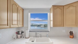 A kitchen with wooden cabinets, a double sink, a window with a view of a body of water and buildings, and a countertop with utensils. - Fort Lauderdale Yacht & Beach Club 411 (Fort Lauderdale)