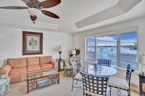 A living room with a sofa, coffee table, and chairs, a dining table with chairs, a ceiling fan, a framed picture, a lamp, and a view of the water with boats.