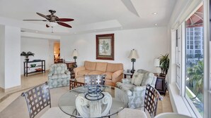 A spacious living room with a ceiling fan, a beige sofa, a glass coffee table, and a view of palm trees through large windows. - Fort Lauderdale Yacht & Beach Club 411 (Fort Lauderdale)