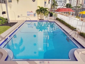 Ein rechteckiger Swimmingpool mit einer Tiefenmarkierung von 2,4 Metern, umgeben von einem weißen Zaun, Palmen und einem roten Sonnenschirm.