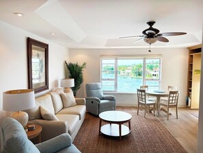 Un salon comprenant un canapé, des fauteuils, une table basse, une table à manger avec des chaises, un ventilateur de plafond et une grande fenêtre donnant sur un plan d’eau et un espace verdoyant.