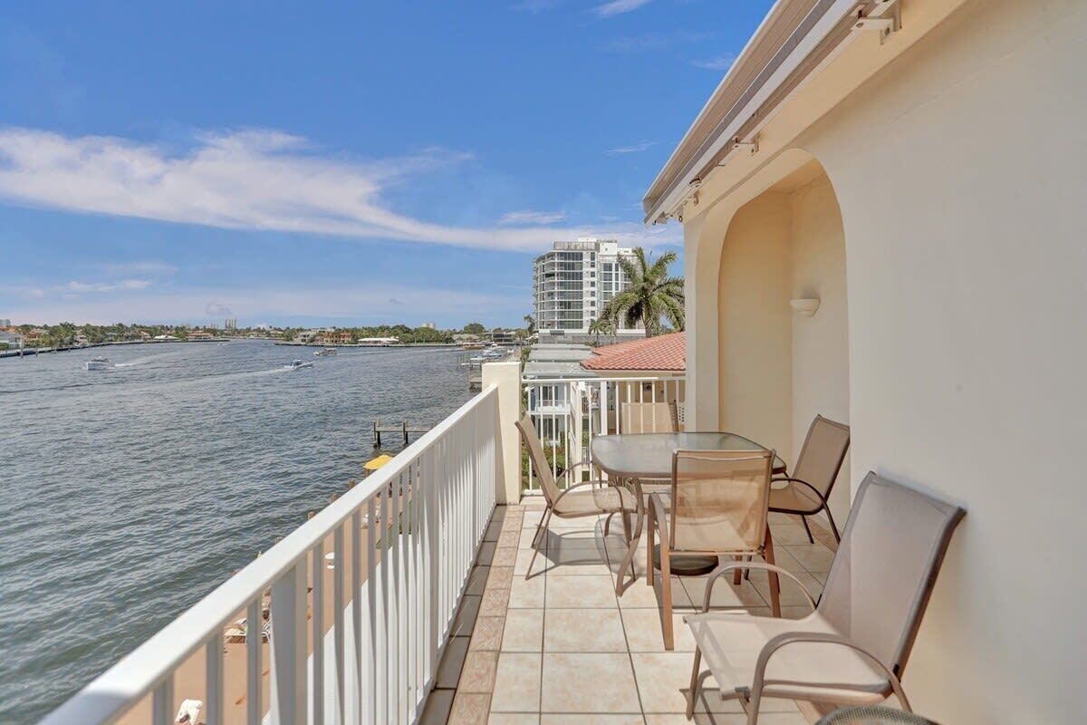 A balcony with a view of the water, a table with chairs, and a building in the background.