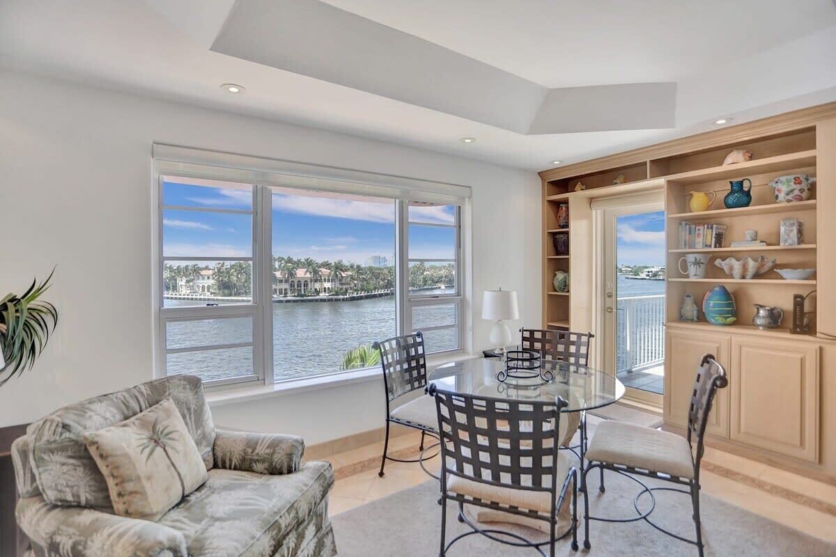 A room with a glass table and chairs, a sofa, a bookshelf, and a view of a body of water.