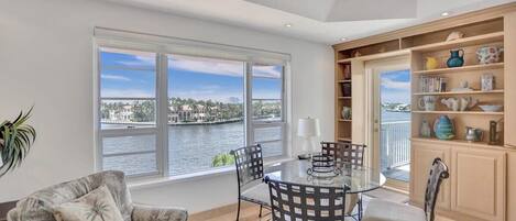 A room with a glass table and chairs, a sofa, a bookshelf, and a view of a body of water.
