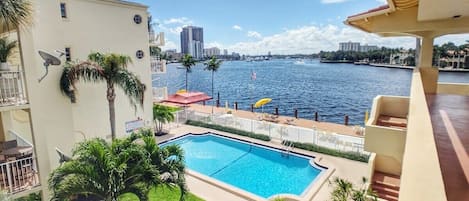 A swimming pool with a view of the water and distant buildings, surrounded by greenery and a balcony with a railing.