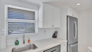 A modern kitchen with a stainless steel refrigerator, a built-in dishwasher, and a window with blinds.