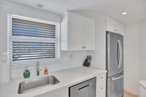 A modern kitchen with white cabinets, a stainless steel refrigerator, a sink with dish soap, and a window with blinds.
