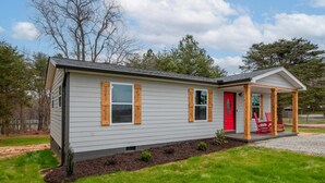 Una casa pequeña de un piso con puerta roja y contraventanas de madera, un porche cubierto con sillas rojas y un césped bien cuidado.