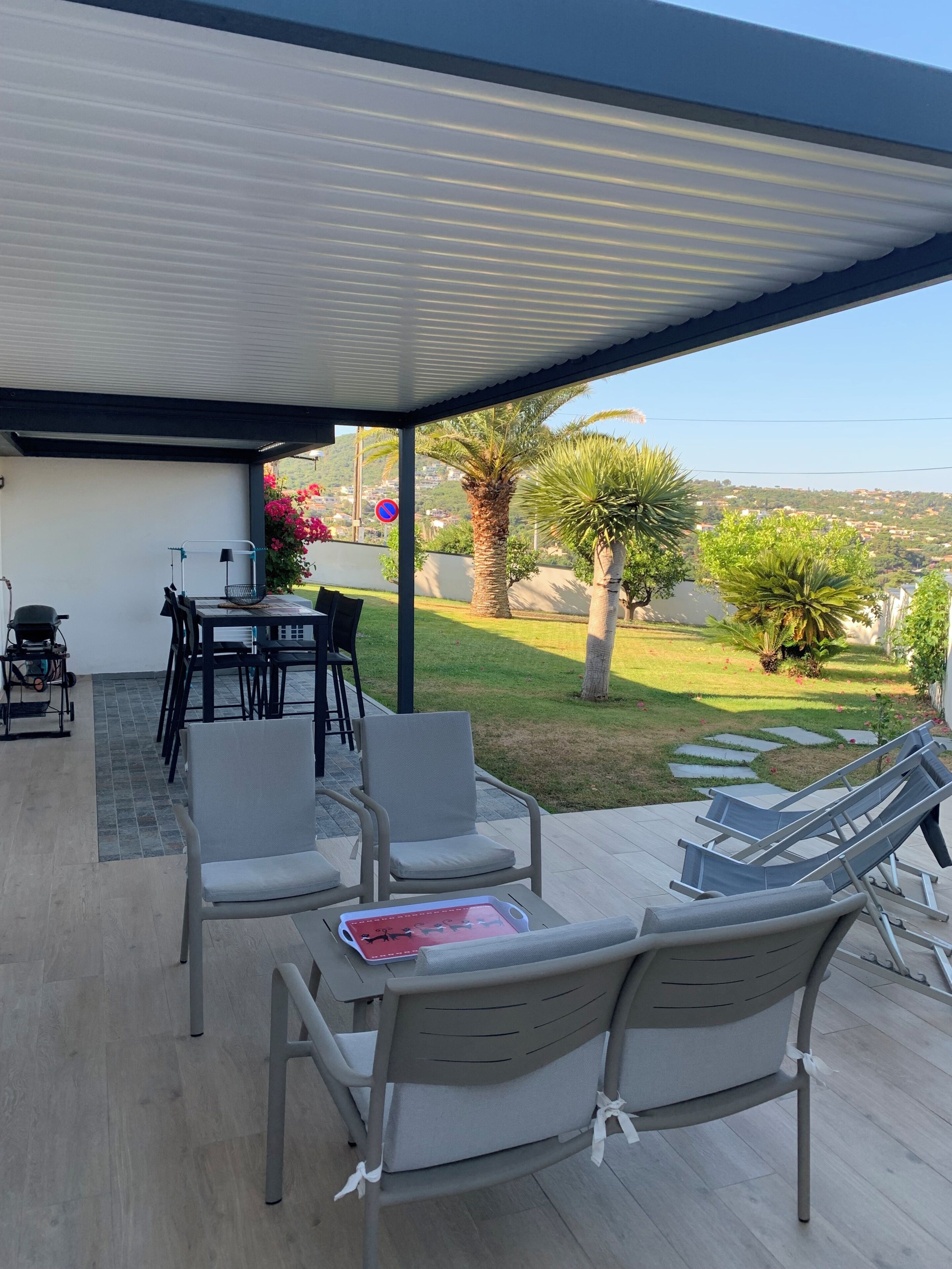 Eine überdachte Terrasse im Freien mit Sitzgelegenheiten, einem Tisch und Blick auf einen angelegten Garten und ferne Hügel.