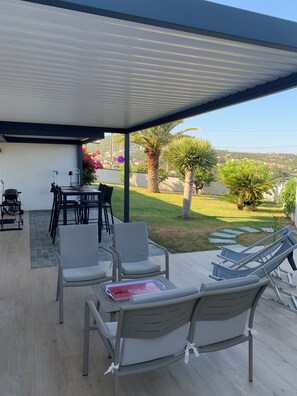 Un patio cubierto al aire libre con asientos, una mesa y vistas a un jardín ajardinado y colinas distantes.