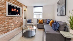 Un salon moderne avec un mur en briques, un canapé gris agrémenté de coussins bleus et orange, une table basse en verre et un téléviseur fixé au mur qui affiche le logo « NETFLIX ».