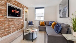 A modern living room with a brick wall, a grey sofa, and a flat-screen TV mounted on the wall displaying 'NETFLIX'. - York Luxury Apt: Bishopthorpe Rd 2, Sleeps 6 (York)