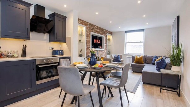 A modern kitchen with dark cabinets, a built-in oven, and a dining area with a round table and chairs. There is a large screen displaying the Netflix logo on the wall. - York Luxury Apt: Bishopthorpe Rd 2, Sleeps 6 (York)