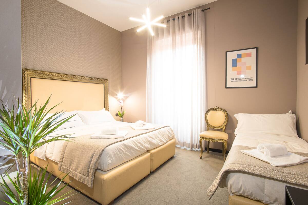 Ein Hotelzimmer mit einem großen Bett, einem kleineren Bett, einem Sessel und einer Topfpflanze.