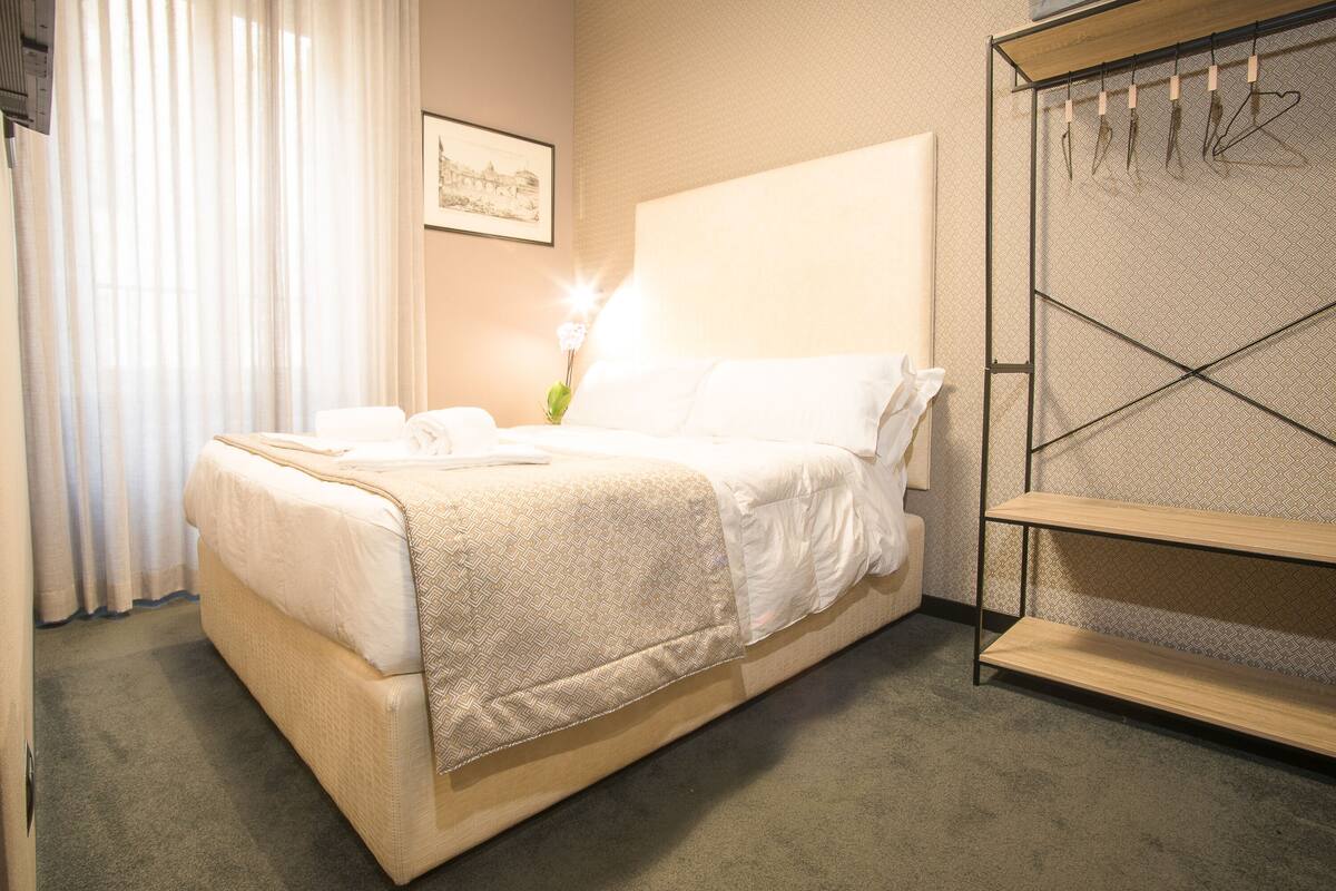 Ein ordentlich bezogenes Bett mit weißer Decke und Kissen, ein Nachttisch mit Lampe und ein an der Wand befestigtes Regal mit Haken.