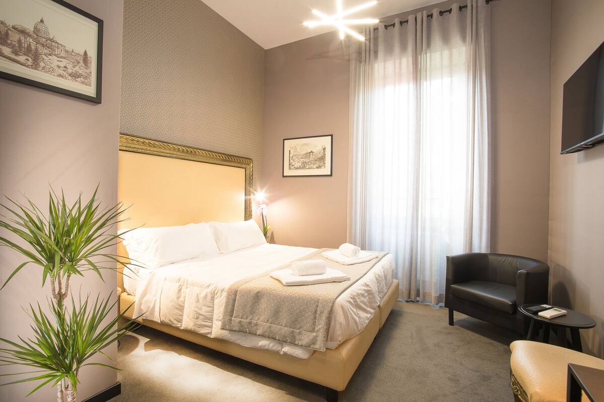 Ein ordentlich eingerichtetes Hotelzimmer mit Bett, Stuhl, kleinem Tisch und einer Zimmerpflanze.