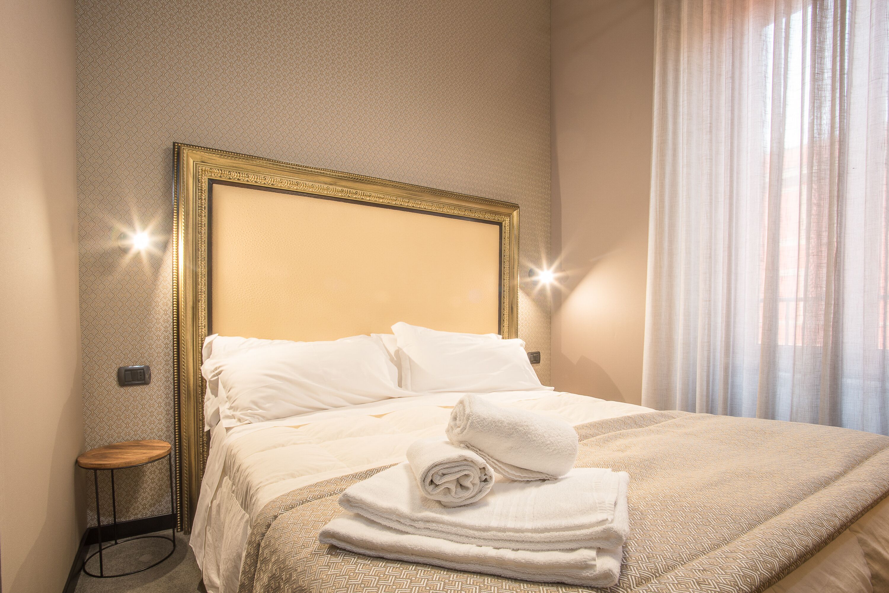 Una cama bien hecha con sábanas blancas y una toalla enrollada, una mesita de noche con una lámpara, y una lámpara de pared.