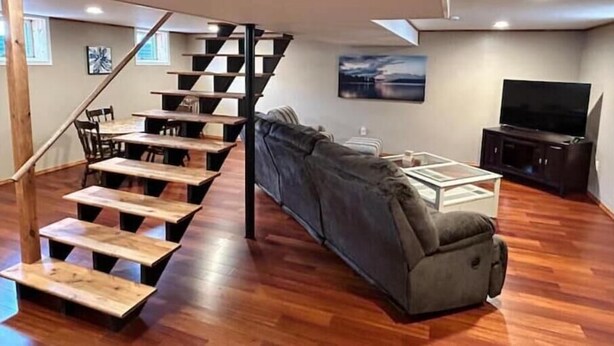A modern living room with a wooden staircase, a gray sofa, a flat-screen TV, and a dining area with wooden tables and chairs. - Golf view! (Red Wing)