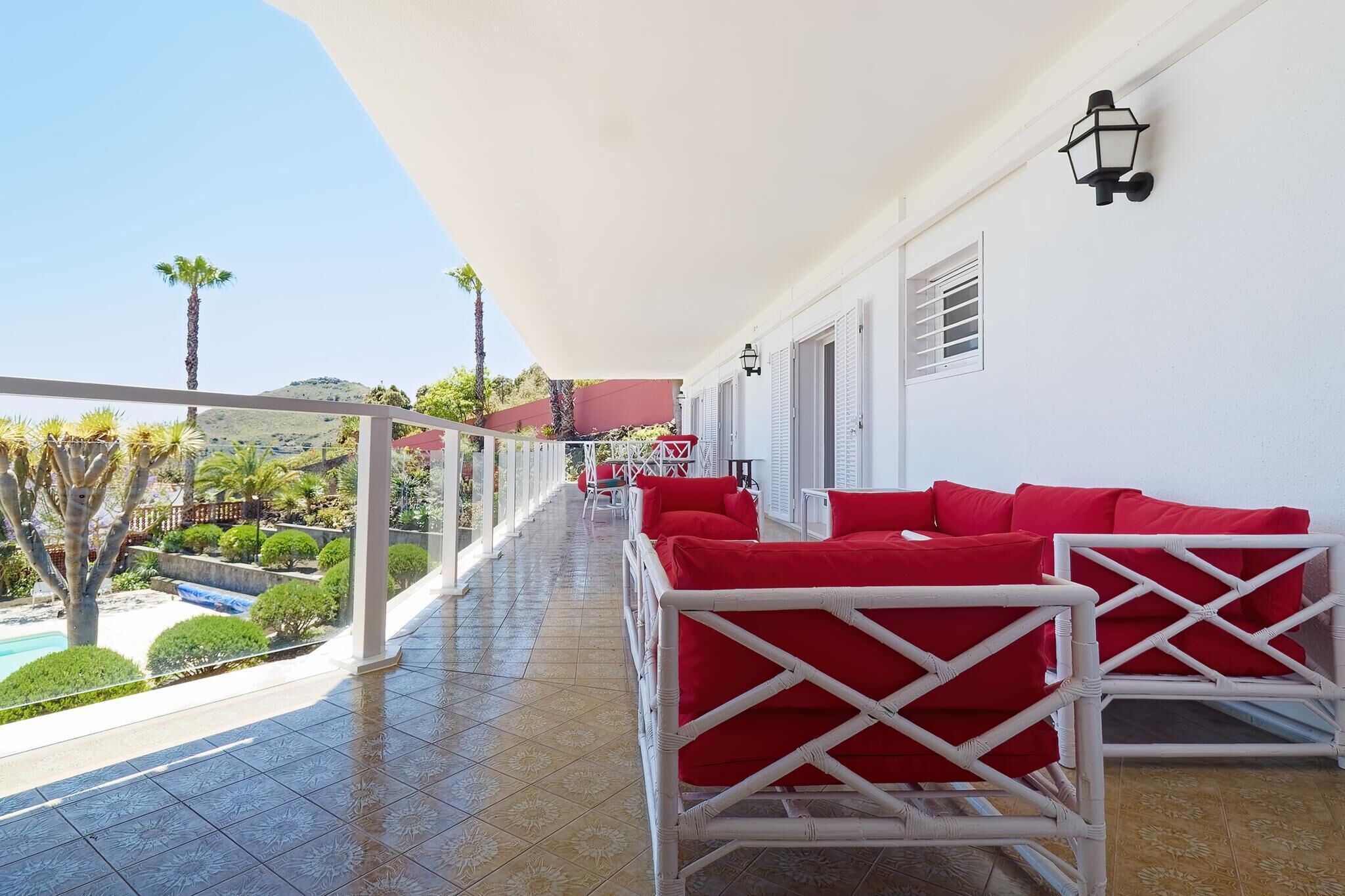 Un balcon doté de meubles en osier blanc et de coussins rouges, offrant une vue sur une piscine et un jardin.