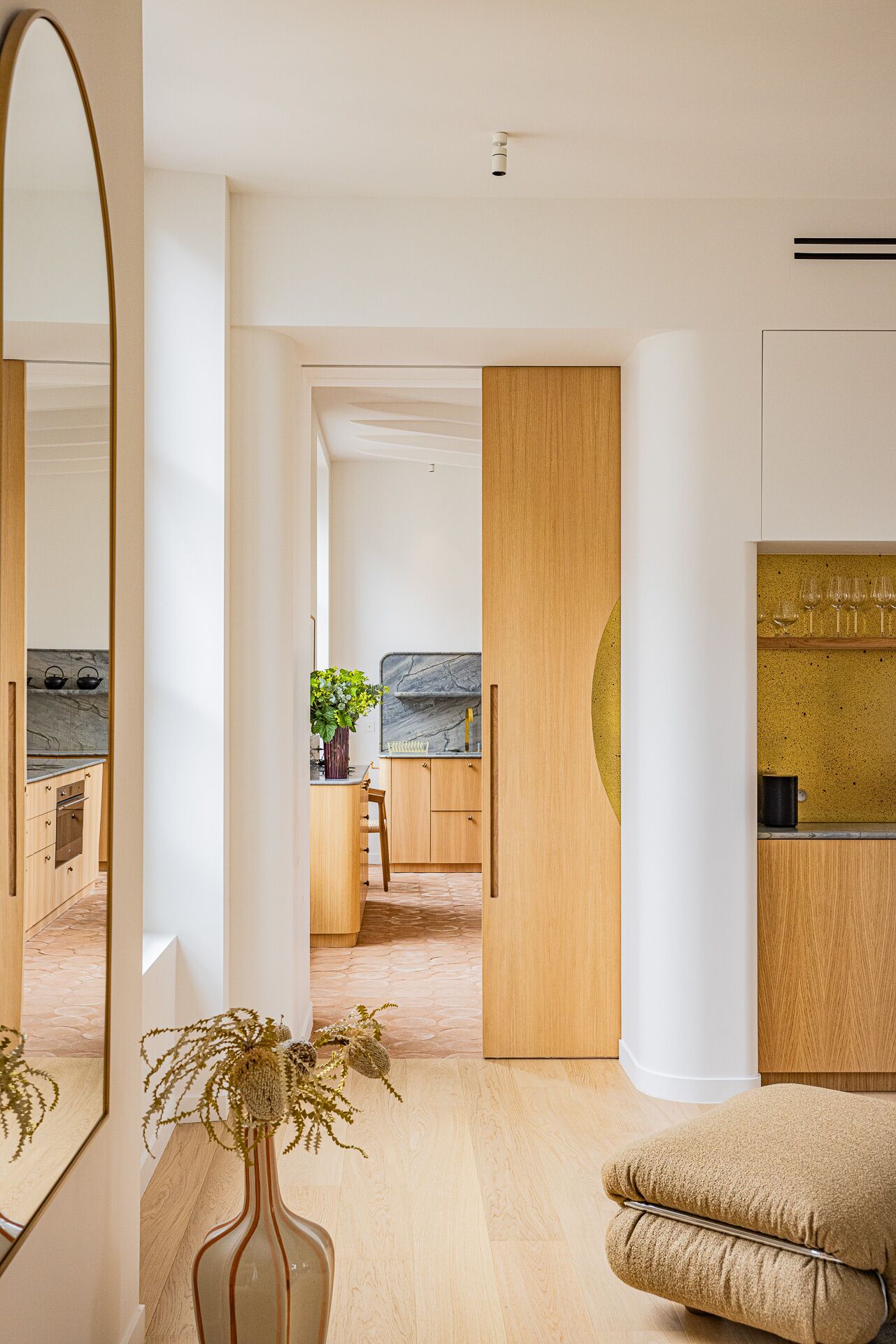 Un interior moderno con zona de comedor de madera, cocina con salpicadero de mármol y un espejo grande que refleja el ambiente.