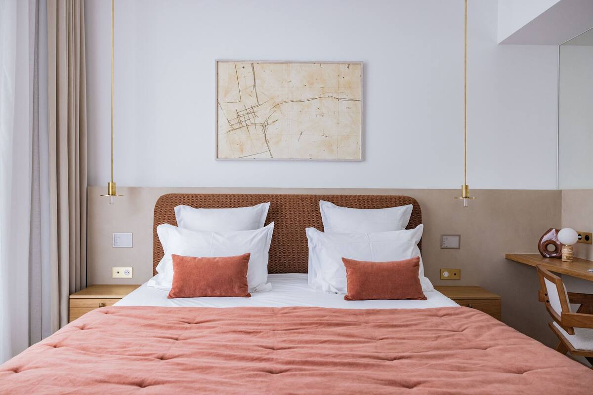 Una cama bien tendida con ropa de cama blanca y marrón, una obra de arte abstracta en la pared y una mesita de noche de madera con una lámpara.