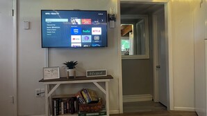 A living room with a flat-screen TV mounted on the wall, a small table with a plant, and a bookshelf.