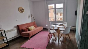 Un salón con un sofá rosa, una mesa redonda con sillas, un radiador y una ventana con vistas a edificios.