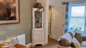 A dining area with a wooden table set for four, a tall white cabinet with glass doors, and a framed deer painting on the wall. - Berry View - Idyllic cosy cottage on berry farm (Rattray)