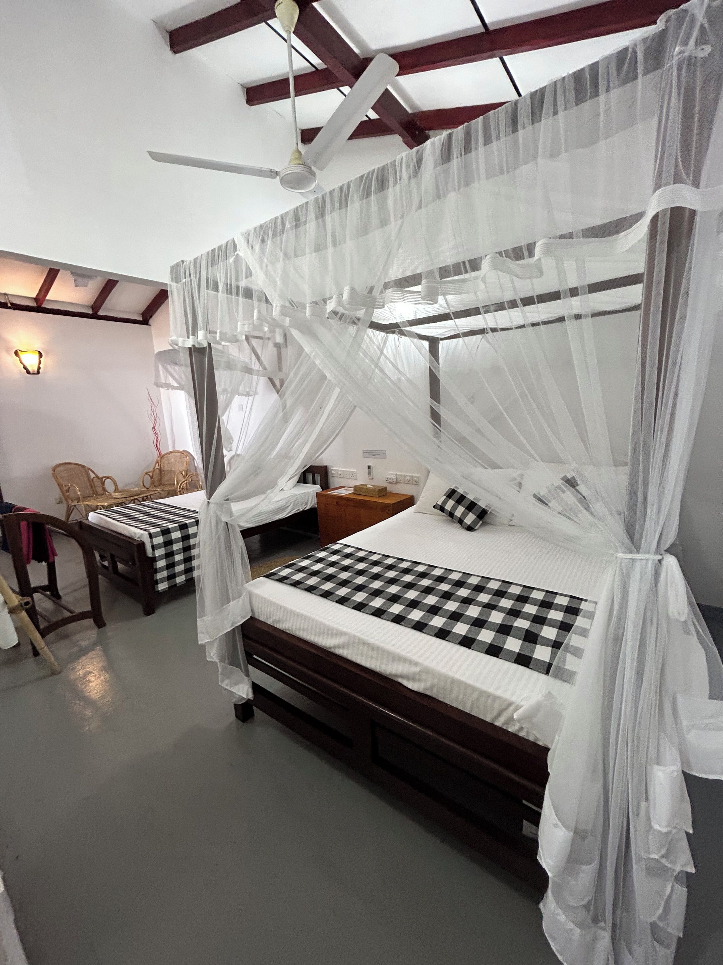 A room with two beds, each with a canopy, a checkered bedspread, and a bedside table with a lamp. There is a chair and a small table with a box on it.