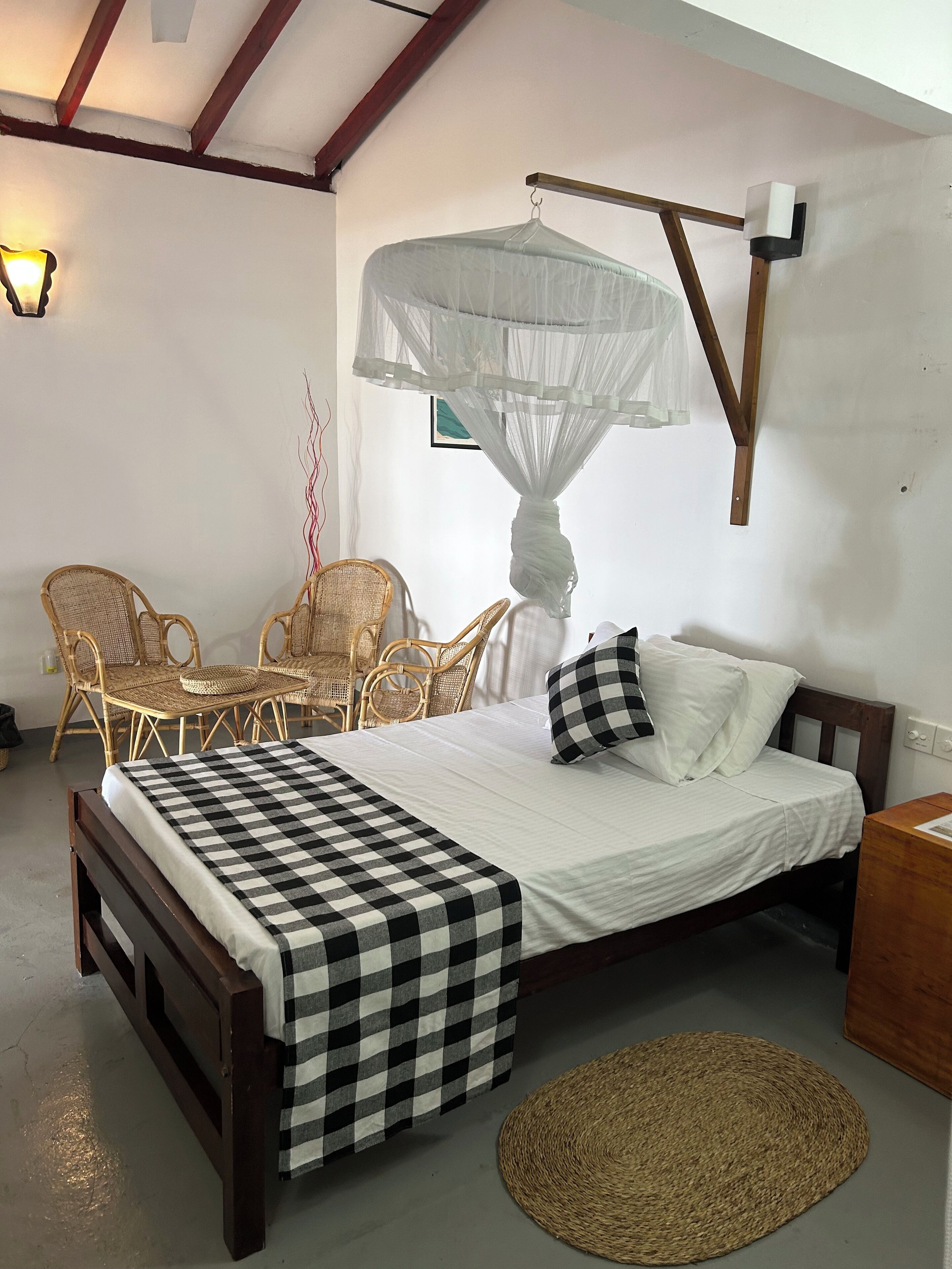 A bedroom with a bed, checkered bedspread, a mosquito net, wicker furniture, and a woven mat.
