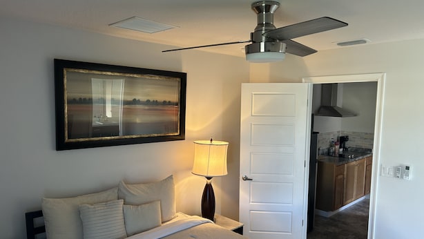 Ein Hotelzimmer mit Bett, Nachttisch, Lampe, Deckenventilator, einem Gemälde und Blick in die Küche.