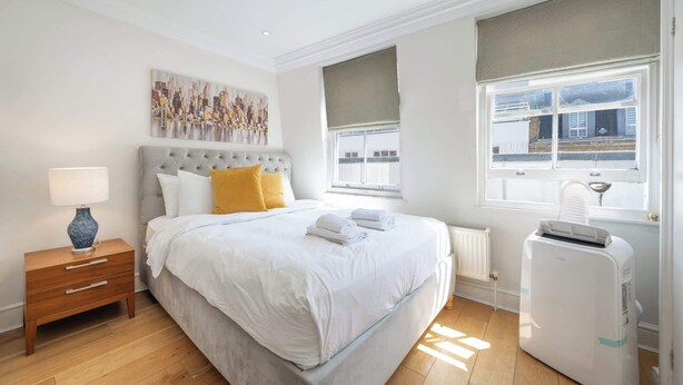A bedroom with a bed, a nightstand, a radiator, a window, and a wall-mounted artwork. - Cozy 2-Bedroom Loft next to Park Lane (London)